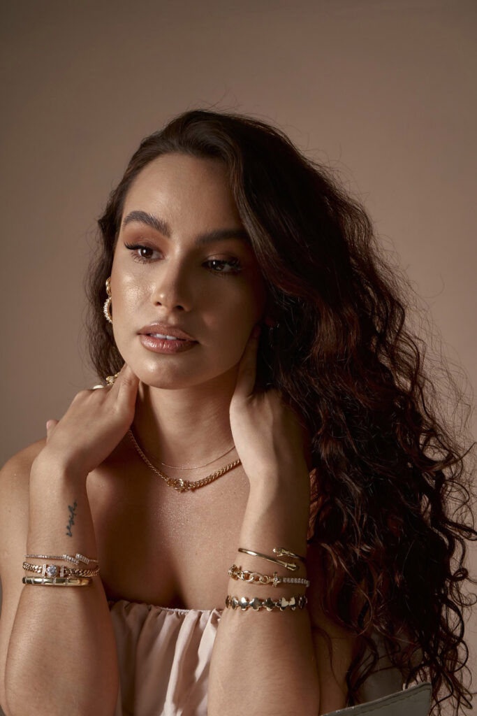 Abril Escalante a model with long wavy brown hair is captured in a portrait against a muted beige background. She wears a delicate gold necklace and multiple gold bracelets on her wrists with one of the bracelet bands bearing a tattoo. Her makeup is subtle emphasizing her eyes and lips and she has a soft neutral expression on her face.
